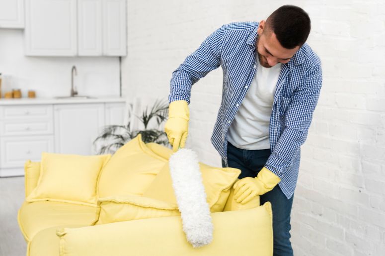 Manutenção de estofados com limpeza de sofá amarelo, feita com espanador para remover poeira de almofadas e encosto em ambiente residencial