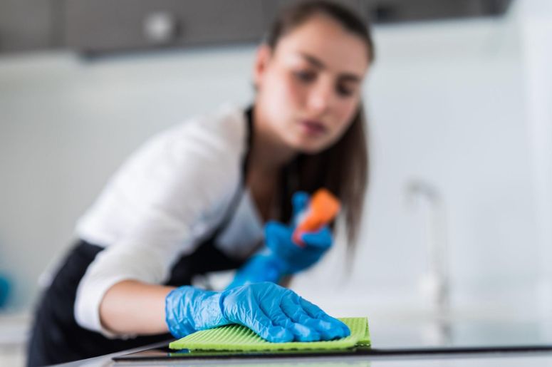 Mulher realizando limpeza residencial com luvas azuis, pano verde e borrifador, higienizando a bancada de uma cozinha em serviço de limpeza profissional