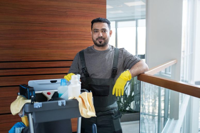 Profissional de limpeza com luvas amarelas e carrinho com produtos, panos e equipamentos para serviço de limpeza profissional em ambiente corporativo