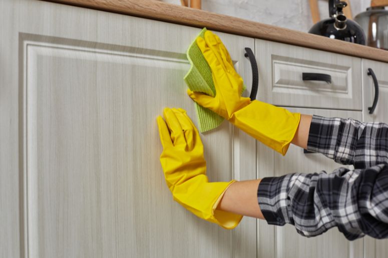 Mãos com luvas amarelas limpando armário de cozinha com pano verde, ação importante para a conservação de móveis modulados
