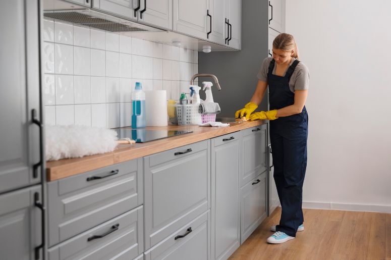 Profissional realizando a conservação de móveis modulados na cozinha, com luvas amarelas, pano de limpeza e produtos sobre a bancada