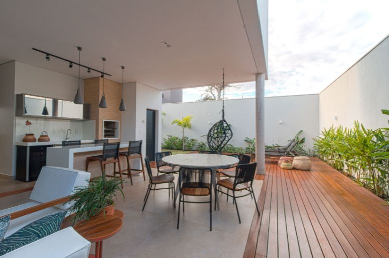 Varanda gourmet com mesa de jantar, bancada com churrasqueira, deck de madeira e área de convivência integrada ao jardim