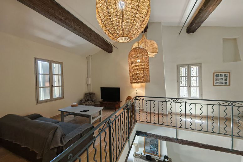 Mezanino com sala de estar no pavimento superior, guarda-corpo metálico, luminárias pendentes de fibra natural, vigas de madeira aparentes e TV no ambiente