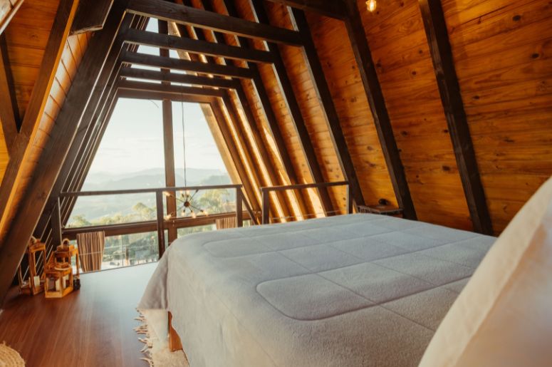 Mezanino com quarto em chalé de madeira, cama de casal, estrutura triangular aparente, grandes janelas de vidro e vista para a paisagem externa