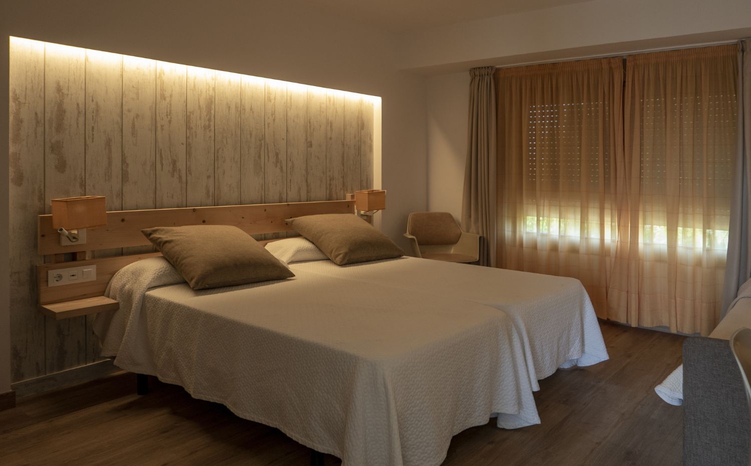 Quarto de hóspedes com cama de casal, cabeceira de madeira, iluminação indireta, cortinas em tons neutros e poltrona ao lado
