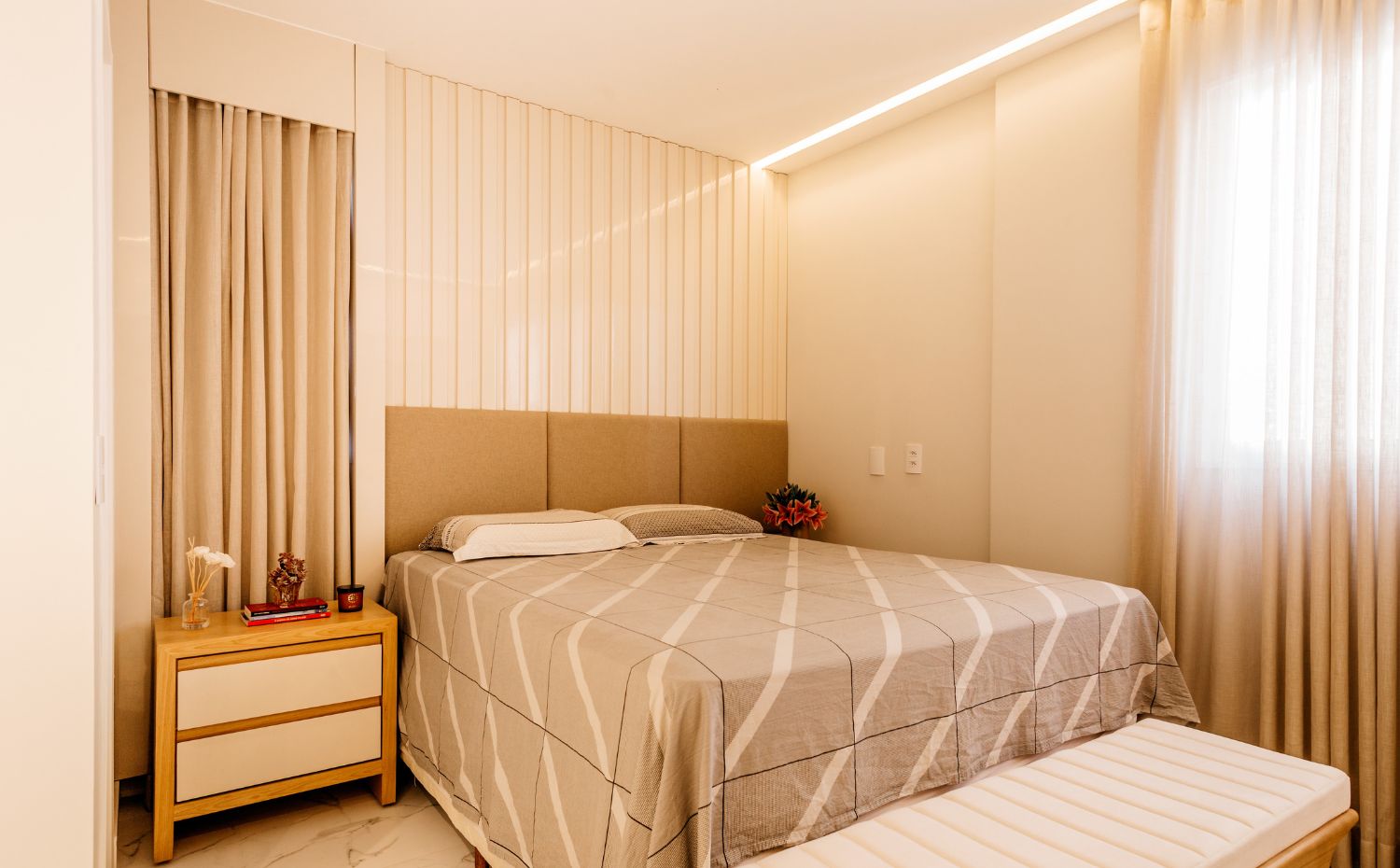 Quarto de casal em tons neutros, com cama, cabeceira estofada, banco aos pés e cortinas claras