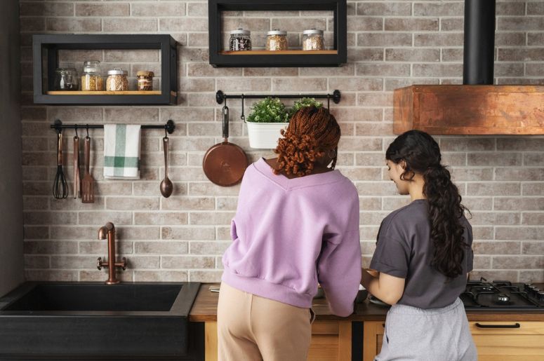 Duas pessoas preparando alimentos em cozinha com prateleiras, utensílios suspensos e acabamento rústico