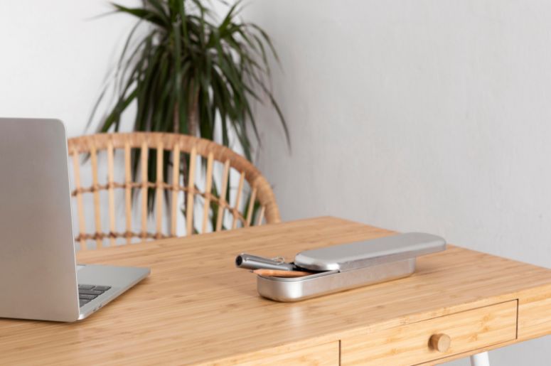 Mesa de madeira com notebook, estojo metálico e cadeira de fibras naturais em ambiente de trabalho com planta ao fundo