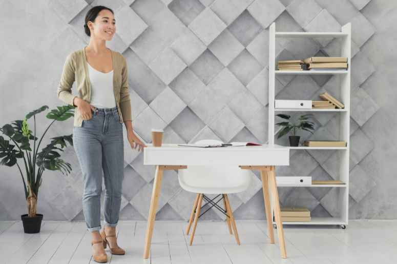 Mulher em home office ao lado de uma mesa branca e uma estante vertical com livros, pastas, caixas e planta