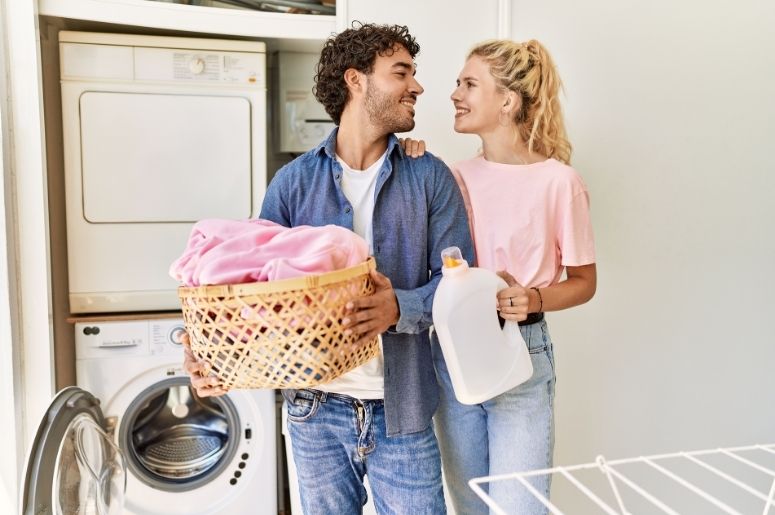Casal organizando a lavanderia, com cesto de roupas, produto de limpeza, máquina de lavar e secadora ao fundo