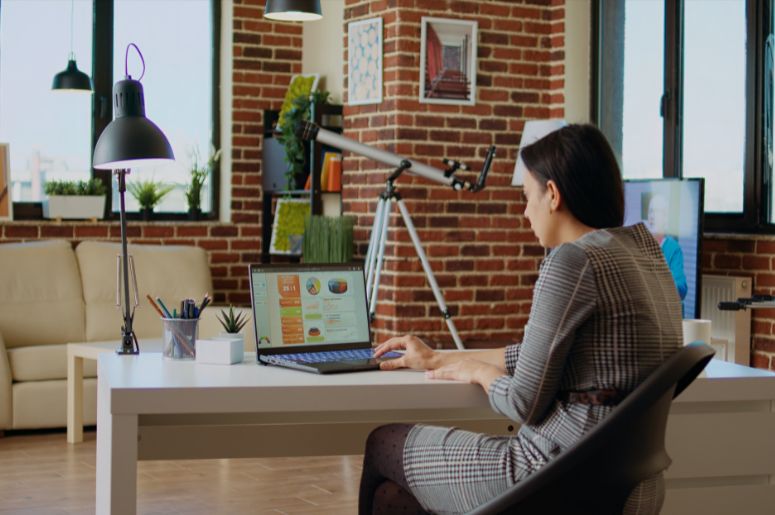 Mulher trabalhando em home office com notebook, mesa ampla e ambiente com estante e iluminação