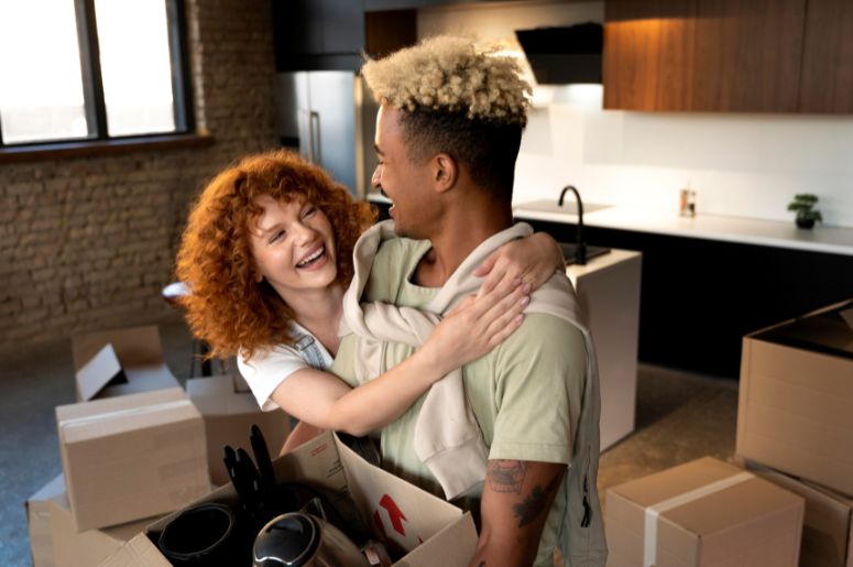 Casal sorrindo abraçado em uma cozinha moderna, cercado por caixas de mudança