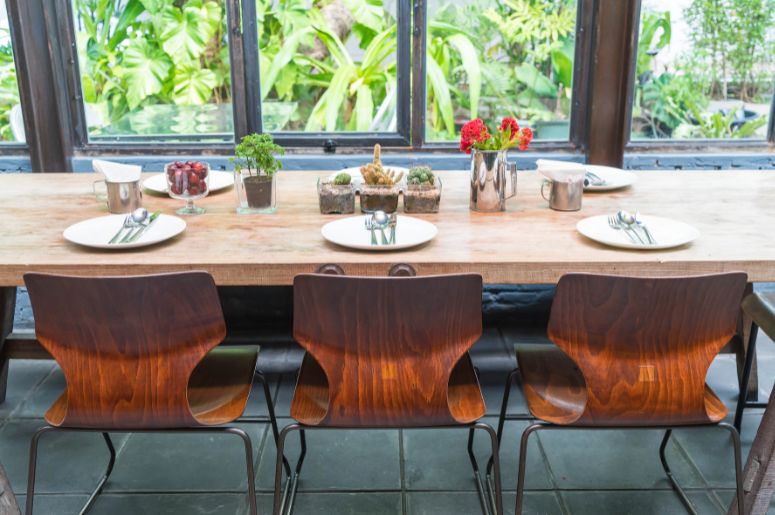 Mesa de jantar retangular de madeira com cadeiras, pratos e plantas decorativas próxima a uma janela