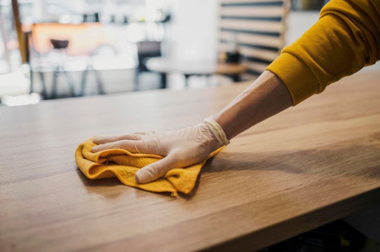 Pessoa limpando uma mesa de madeira com pano de microfibra e luvas de proteção