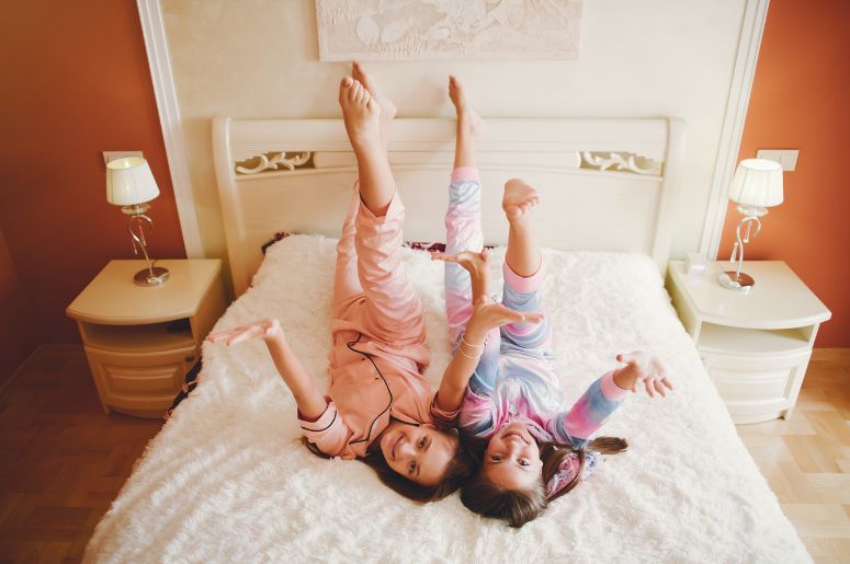 Duas crianças brincando sobre uma cama em quarto infantil com cabeceira e mesas de cabeceira