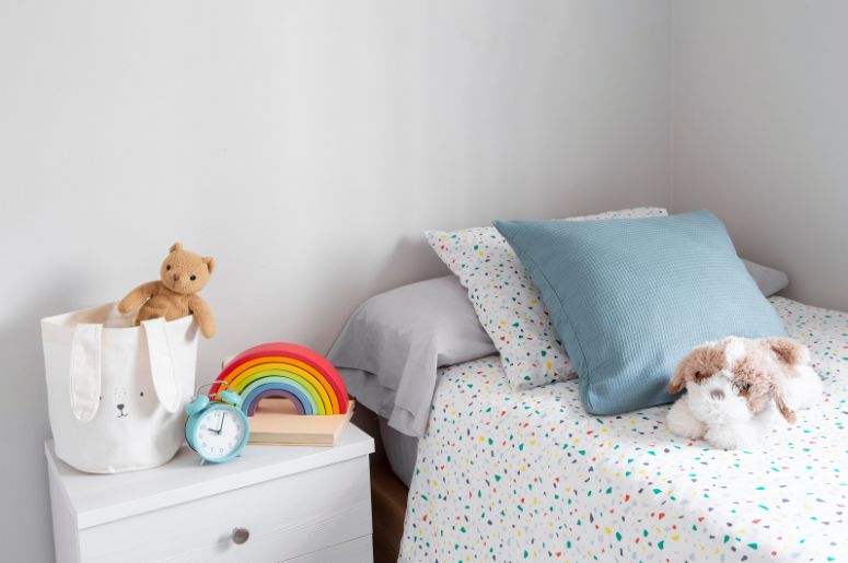 Quarto infantil com cama, mesa de cabeceira branca, brinquedos e objetos decorativos coloridos