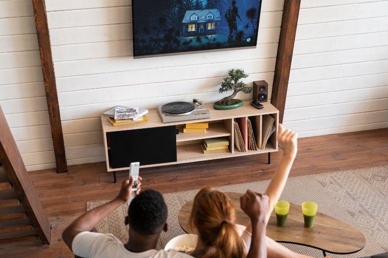 Casal sentado no sofá de costas para a câmera, assistindo televisão em uma sala com piso de madeira, painel de TV suspenso, aparador com toca-discos, livros e plantas decorativas