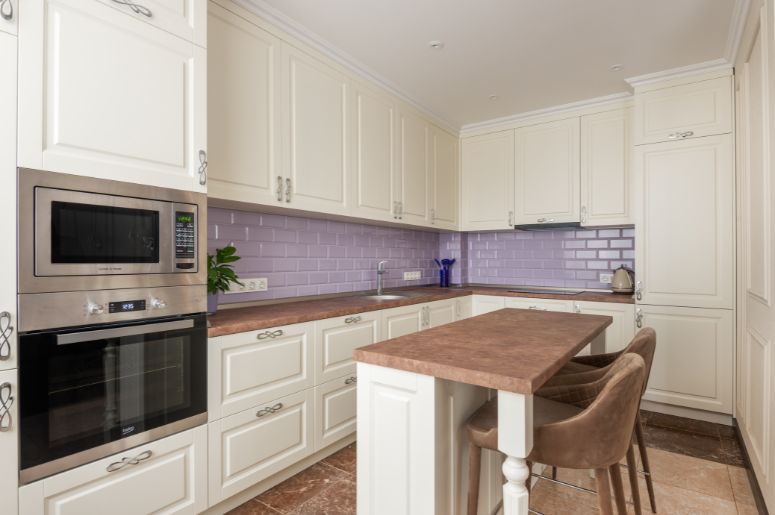 Cozinha provençal com armários brancos, azulejo de metrô lilás e ilha central com banquetas