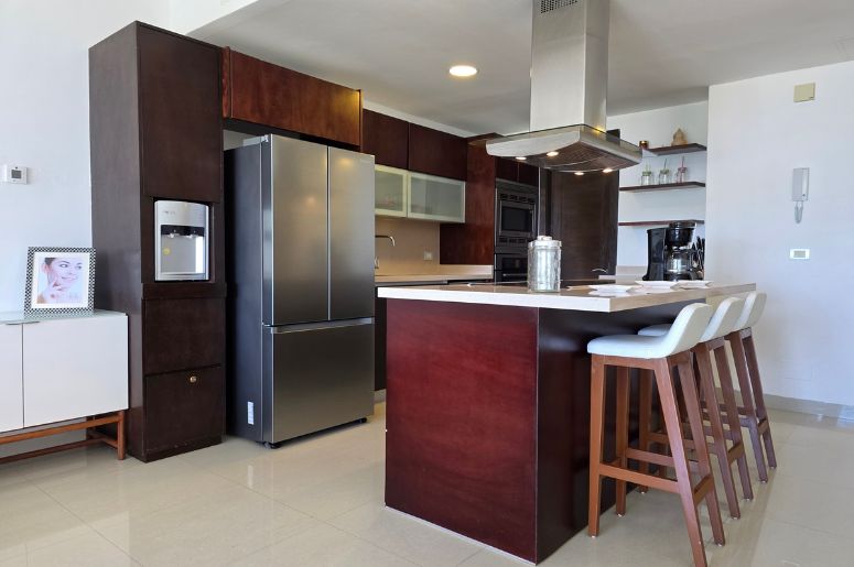 Cozinha americana com ilha em madeira escura, bancada branca e coifa de inox