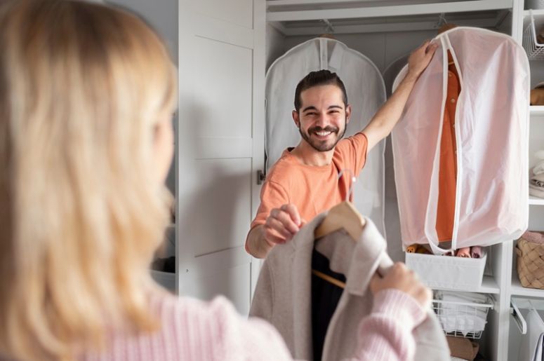 Homem sorrindo entregando um casaco em um cabide para uma mulher em frente ao armário