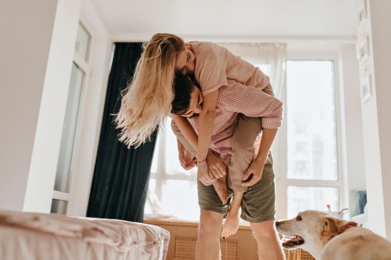Casal se divertindo no quarto perto da cama enquanto organiza a casa, com cachorro ao lado