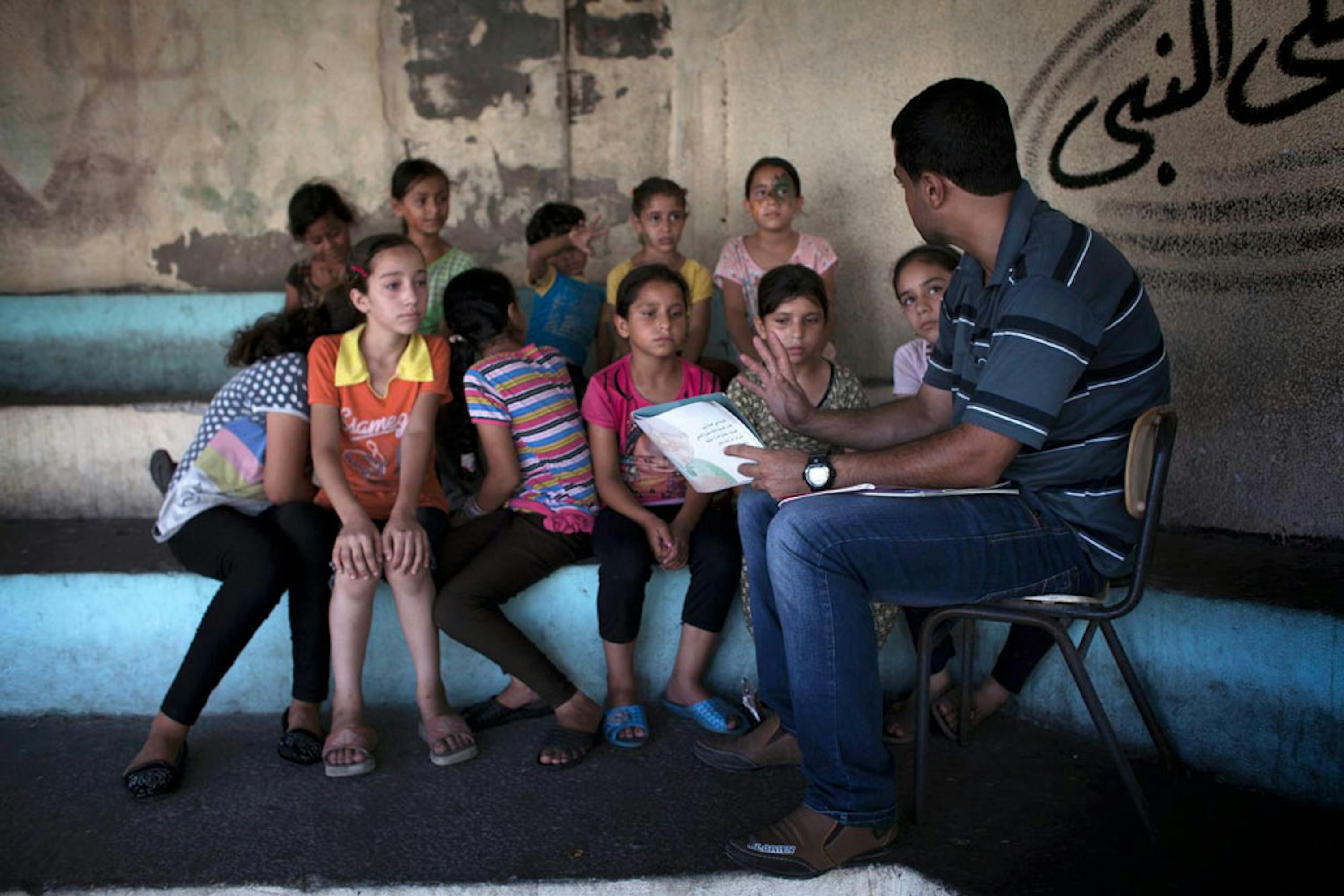 Un volontario legge una storia a un gruppo di bambini sfollati insieme alle loro famiglie in una scuola di Gaza. La foto è stata scattata il 25 agosto, alla vigilia del cessate il fuoco dopo 50 giorni di guerra - ©UNICEF/NYHQ2014-1439/Loulou d'Aki