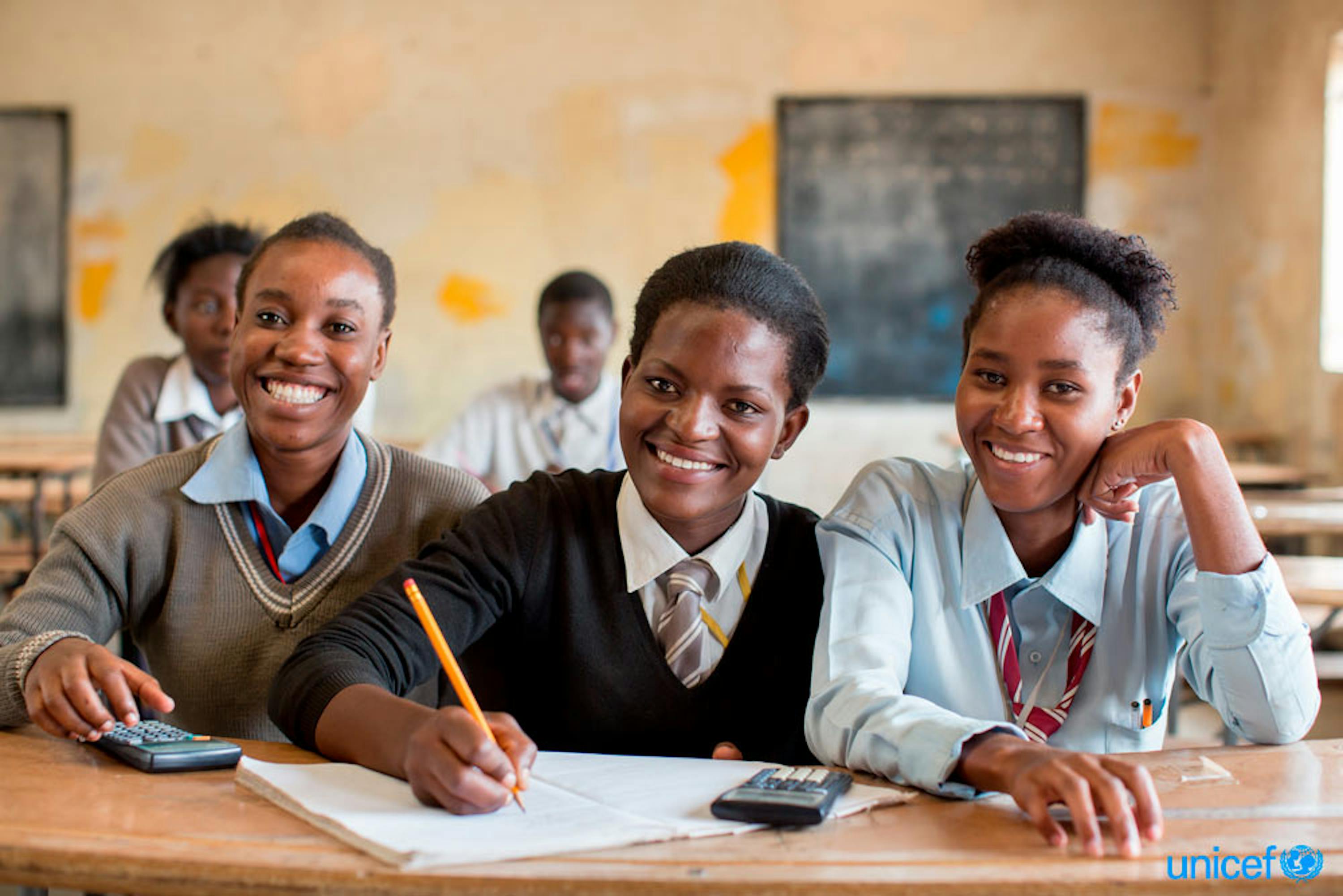 Tre adolescenti, tra cui una che prende appunti, sorridono durante una lezione, nella Kamulanga Secondary School di Lusaka, la capitale - © UNICEF/UN0145551/Schermbrucker