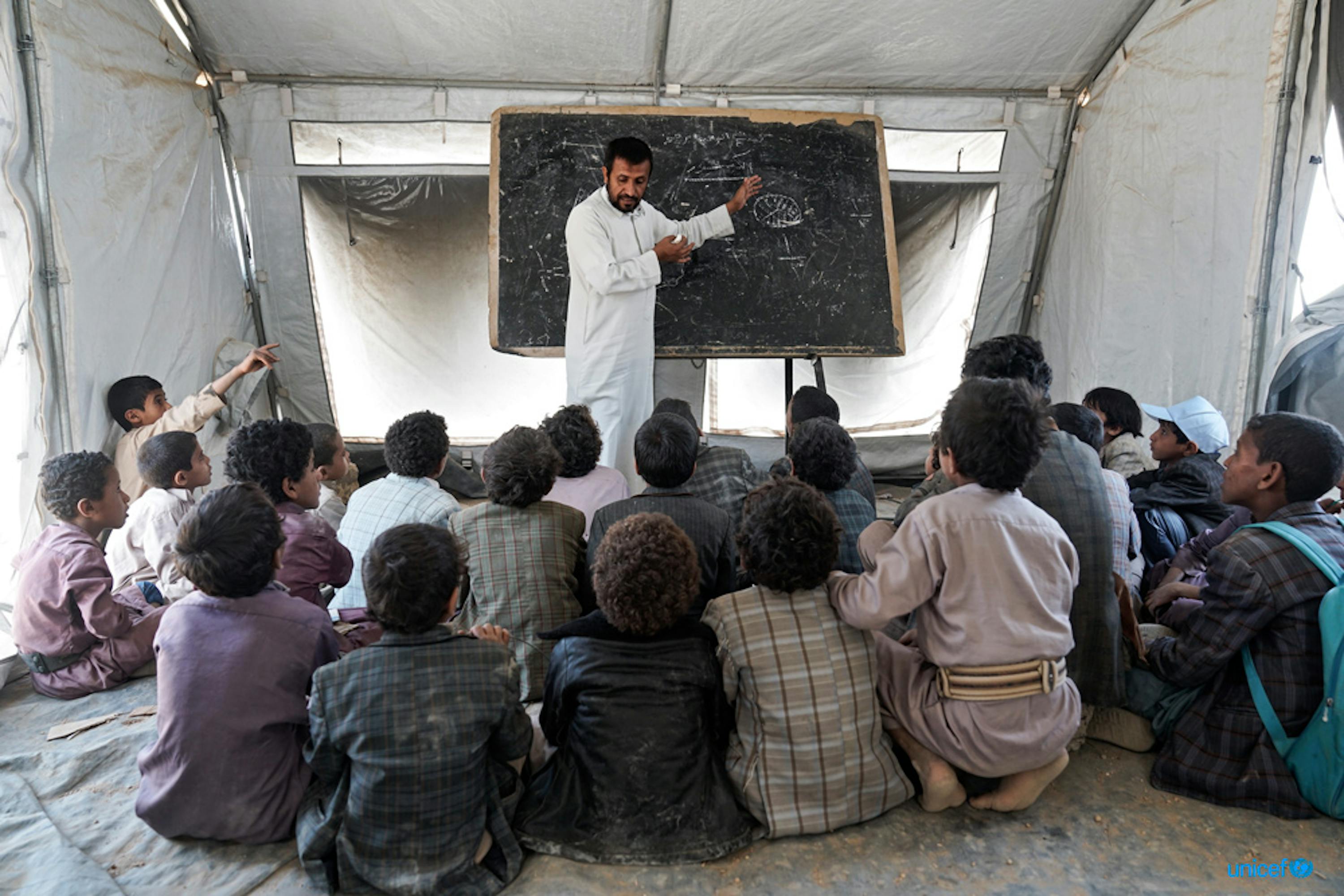 Un gruppo di studenti ascoltano il loro insegnante durante una lezione tenuta in una tenda dell'UNICEF, dopo che la loro scuola Aal Oka B è stata distrutta dai bombardamenti. © UNICEF/UN073956/Clarke for UNOCHA