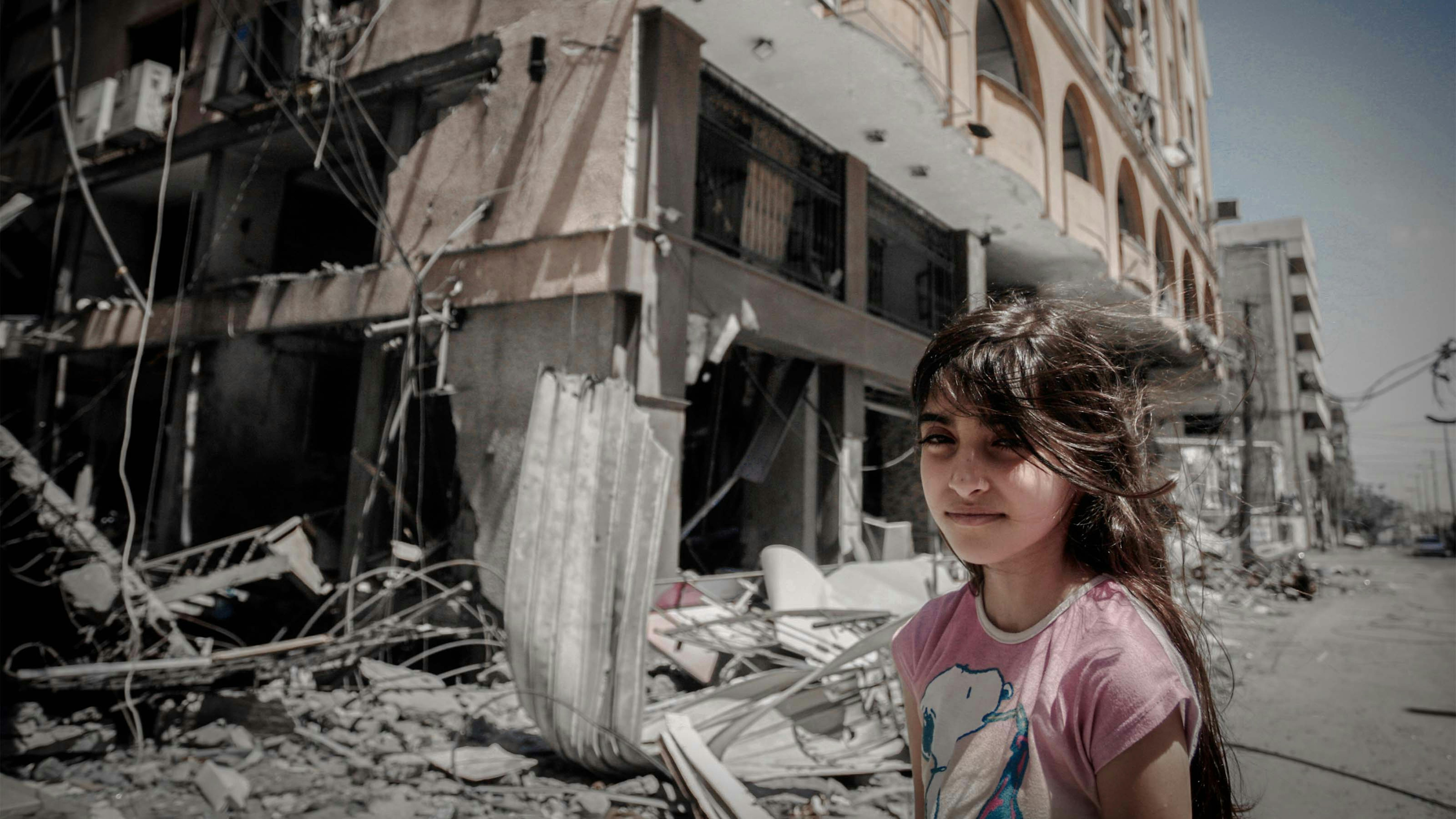 Gaza, 2021. Una ragazza palestinese  guarda in camera mentre sullo sfondo si vede la sua casa distrutta dalle bombe
