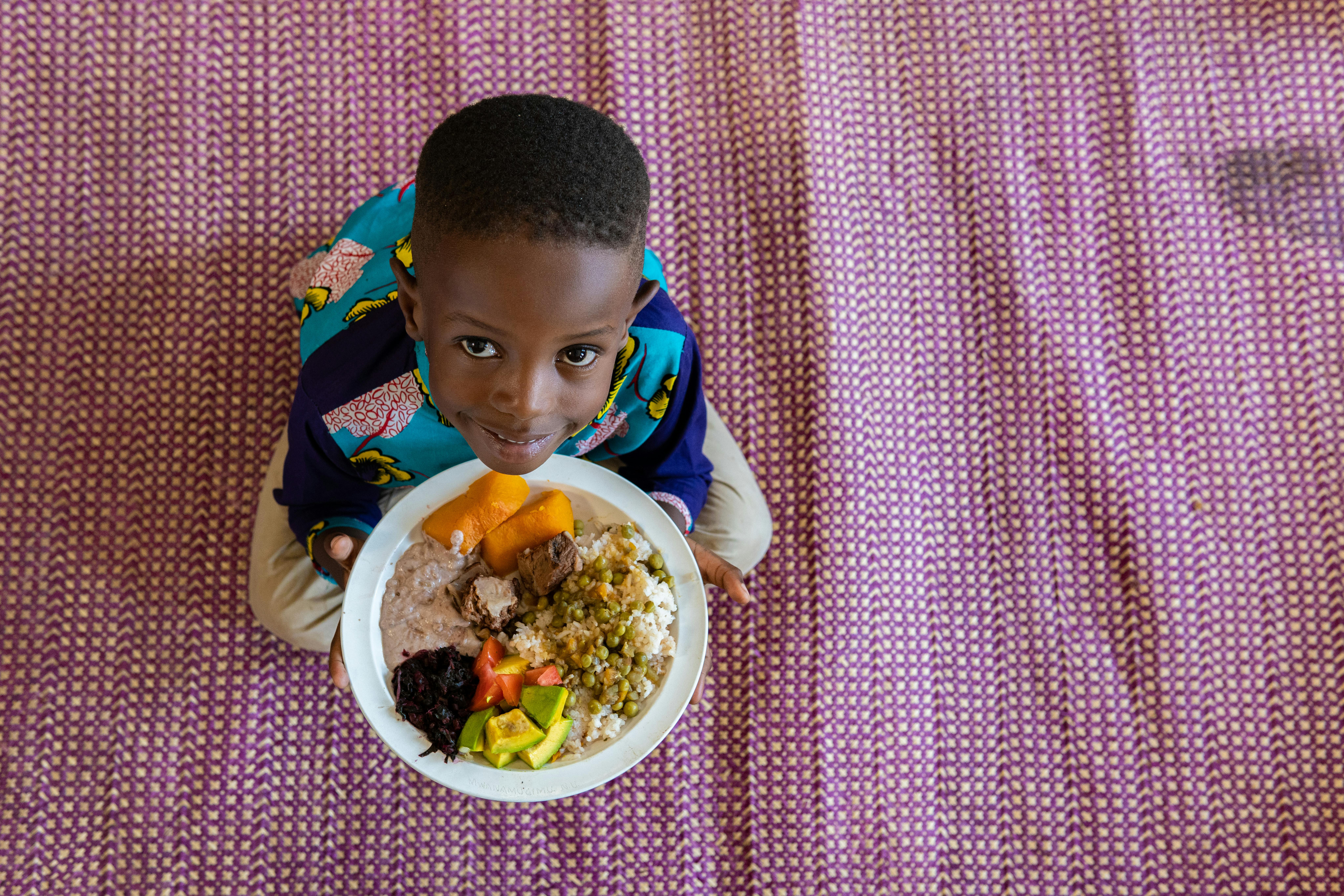 Daniel, 5 anni, vive in Uganda. Tiene in mando il suo pranzo che include zucca, riso, salsa di arachidi, verdure rosse, avocado, piselli e manzo.