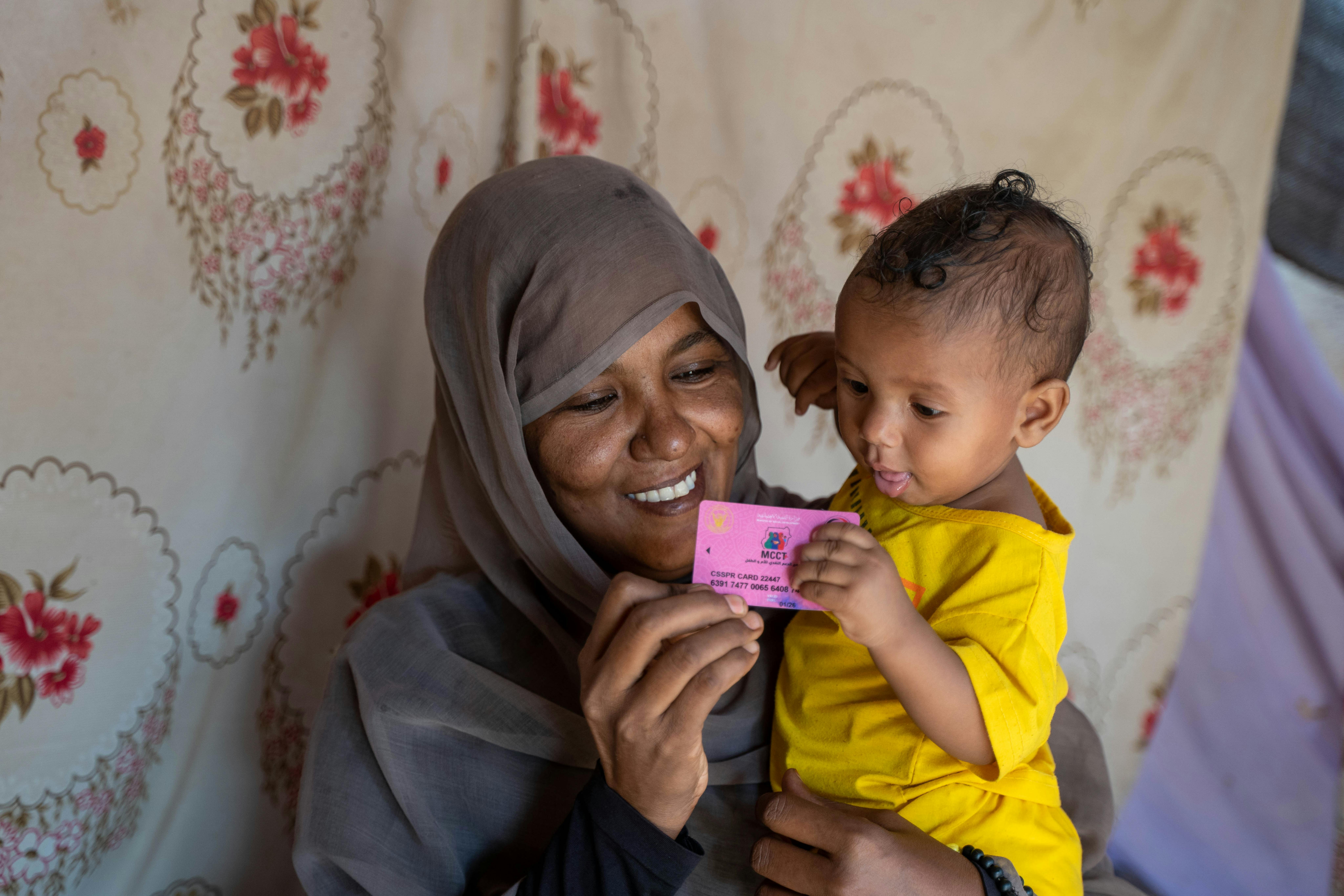 Sabna Mohamed tiene in mano la scheda di registrazione per il programma di trasferimento in contanti per madre e bambino dell'UNICEF (MCCT+), con in braccio sua figlia di 9 mesi, Fatuma, nella loro casa nel Wager Village, North Delta Locality, Kassala State, Sudan.