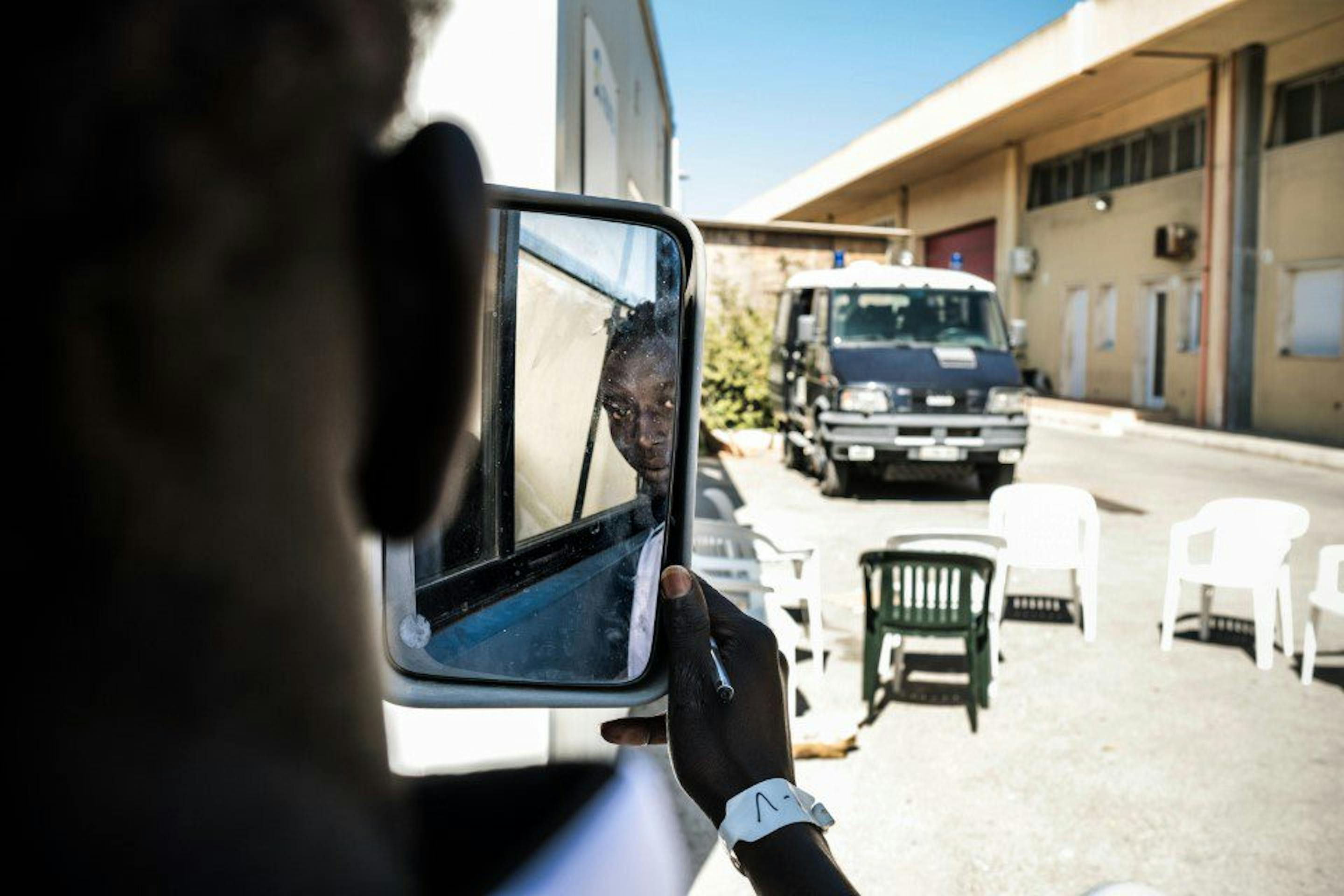 Giovane migrante si guarda allo speccio in macchina, sullo sfondo il furgone della Polizia