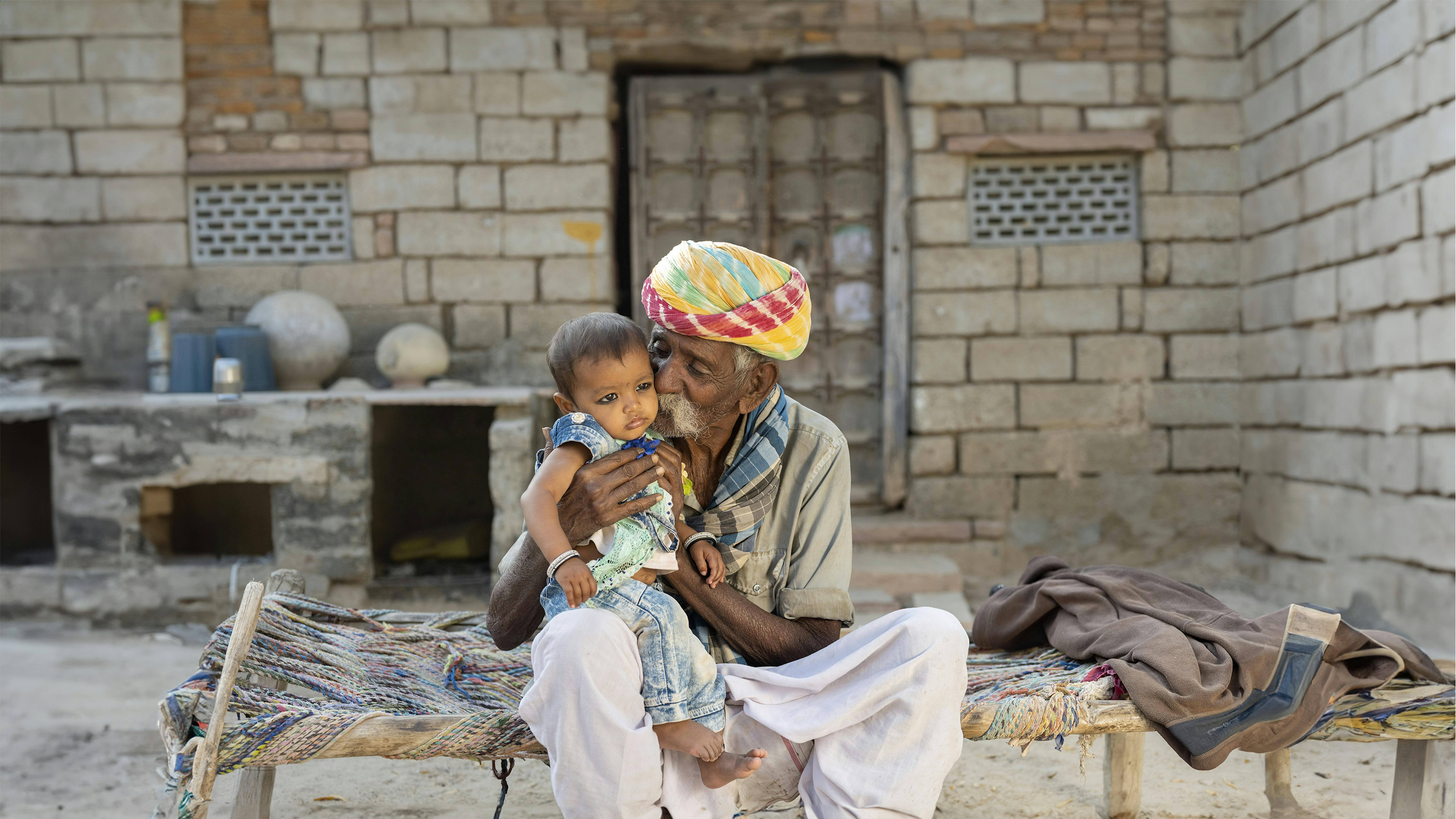 India: Laxmi (9 mesi) e suo nonno sono seduti su un letto khaat indiano. Siamo nel Villaggio di Karnawas, nel Rajasthan