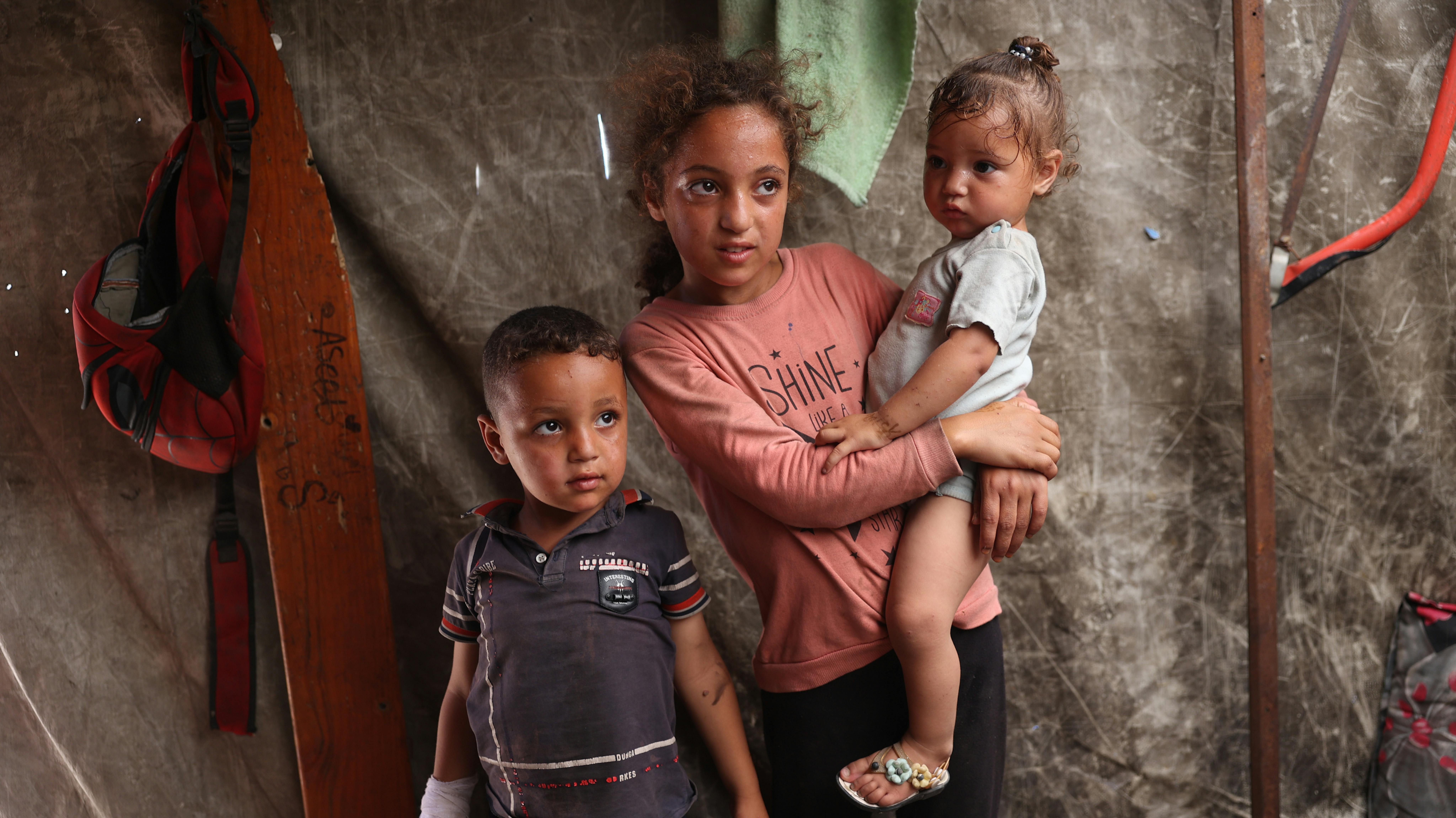 Striscia di Gaza - Una bambina di 11 anni tiene in braccio la sorella di un anno mentre è in piedi con il fratello di 5 anni all'interno della loro tenda a Deir al-Balah