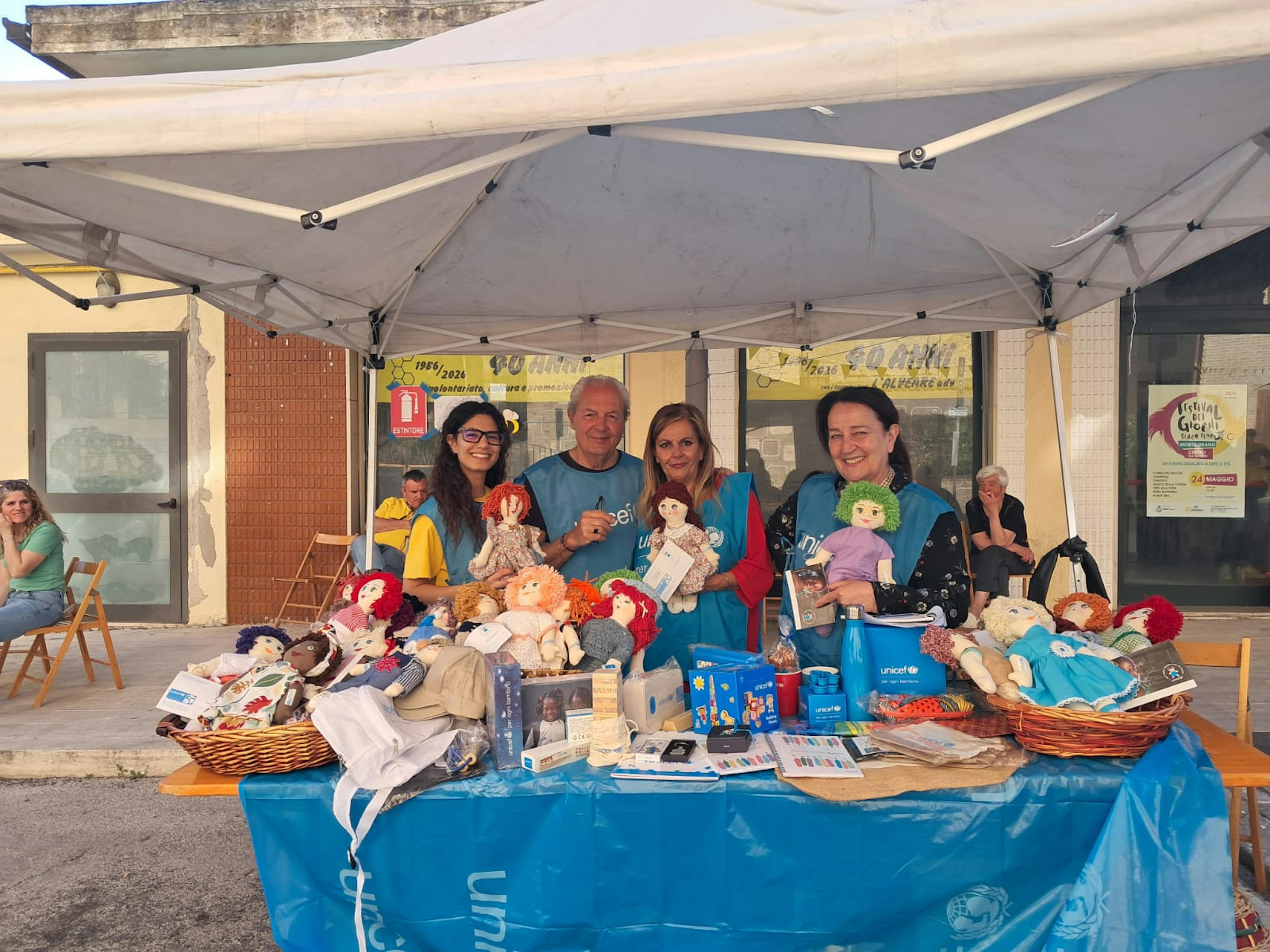 A Monte Urano (Fermo) un gruppo di volontari sorride, mentre mostra le Pigotte e alcuni gadget UNICEF allestiti per la Campagna del Gioco 2026