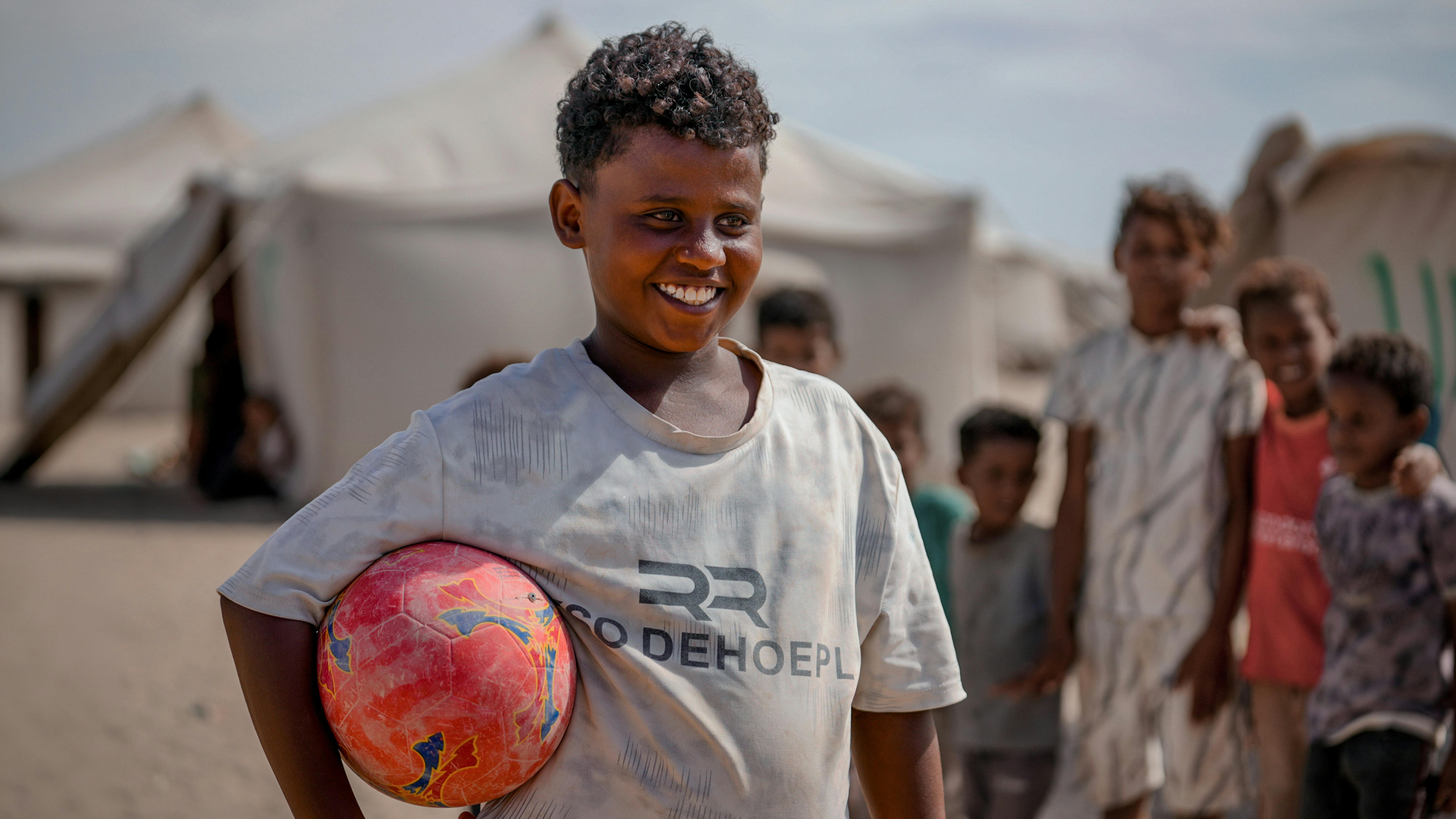 Bassam tiene il suo pallone mentre altri bambini gli corrono accanto nel campo di Al-Firdous, ad Aden.