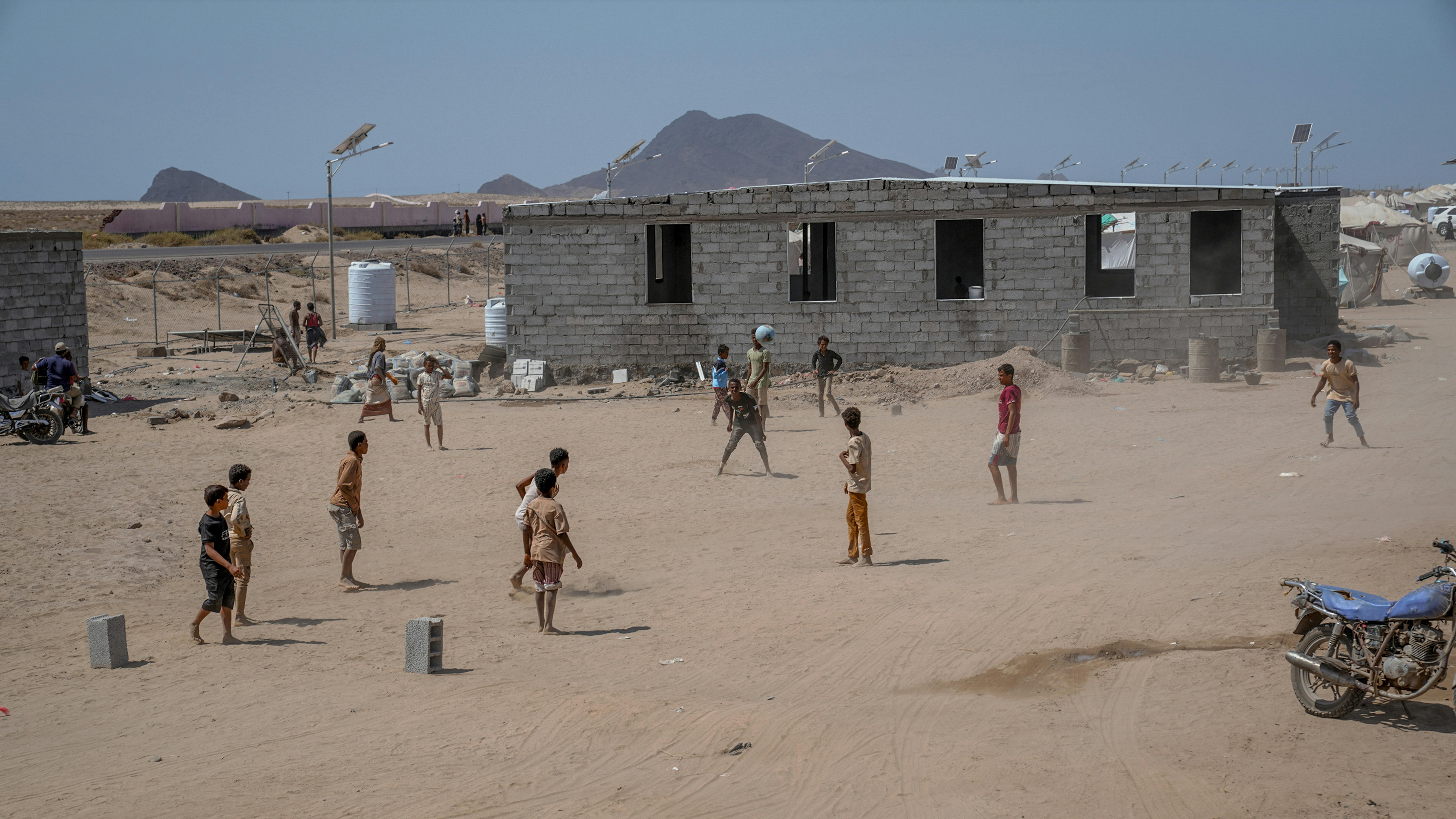 Bambini giocano accanto a uno dei pochi edifici in muratura del campo, con le montagne sullo sfondo. Siamo nel campo sfollati di Al-Firdous, ad Aden in Yemen
