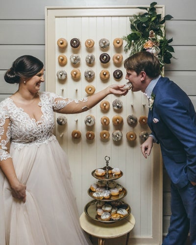Couple feed each other donuts in front of a donut wall