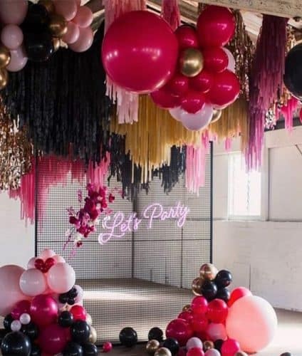 Colourful balloons and garlands hanging from the roof. Metal mesh wall decorated with balloons and a neon sign saying 'lets party'