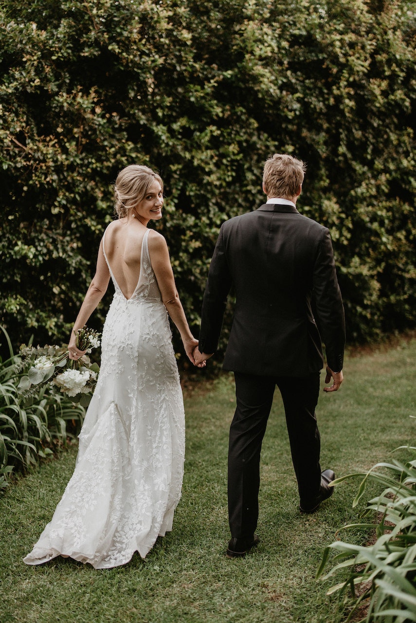 Bride and groom walking through gardens with bride looking over her shoulder 