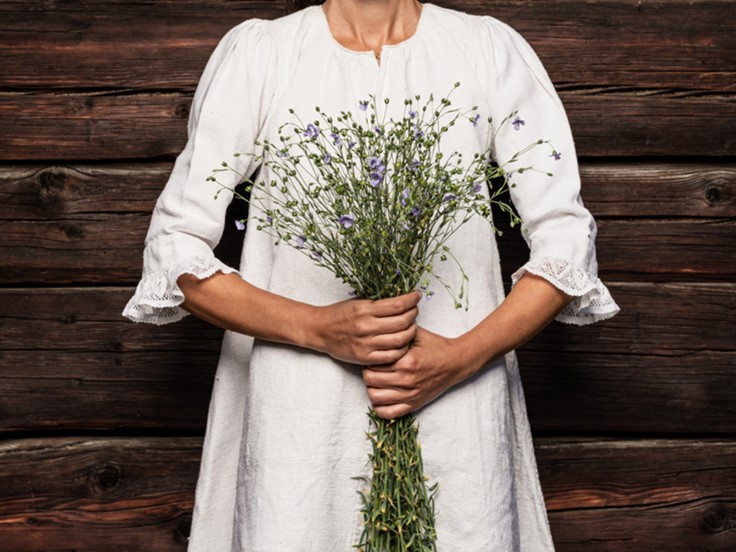 Frau in weissem Leinenkleid hält einen Strauss blau blühender Leinpflanzen