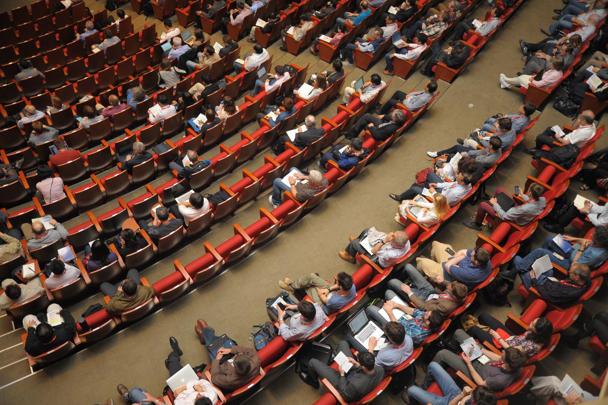 Bird's eye view on a seated conference audience apparently listening to a talk and taking notes on laptops and paper. The speaker is not pictured.