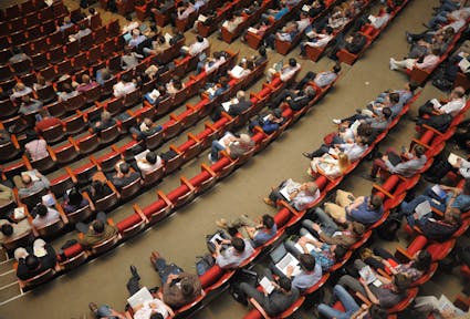 Bird's eye view on a seated conference audience apparently listening to a talk and taking notes on laptops and paper. The speaker is not pictured.