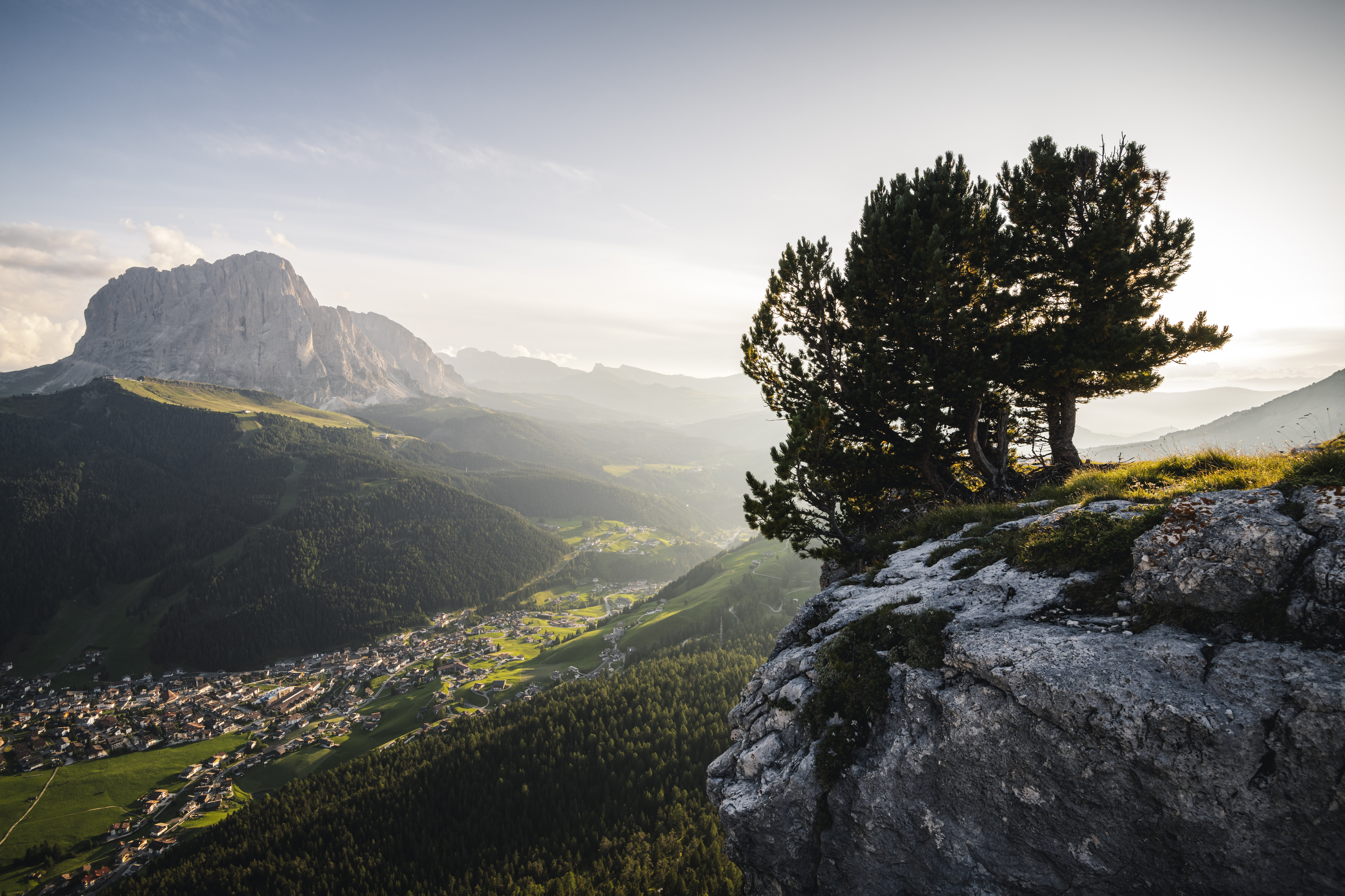Blick über ein leicht nebelverhangenes Grödental. Im Vordergrund ein Baum auf einem Felsbrocken, in der Ebene sind ein Dorf, im Hintergrund ein Berg sichtbar.