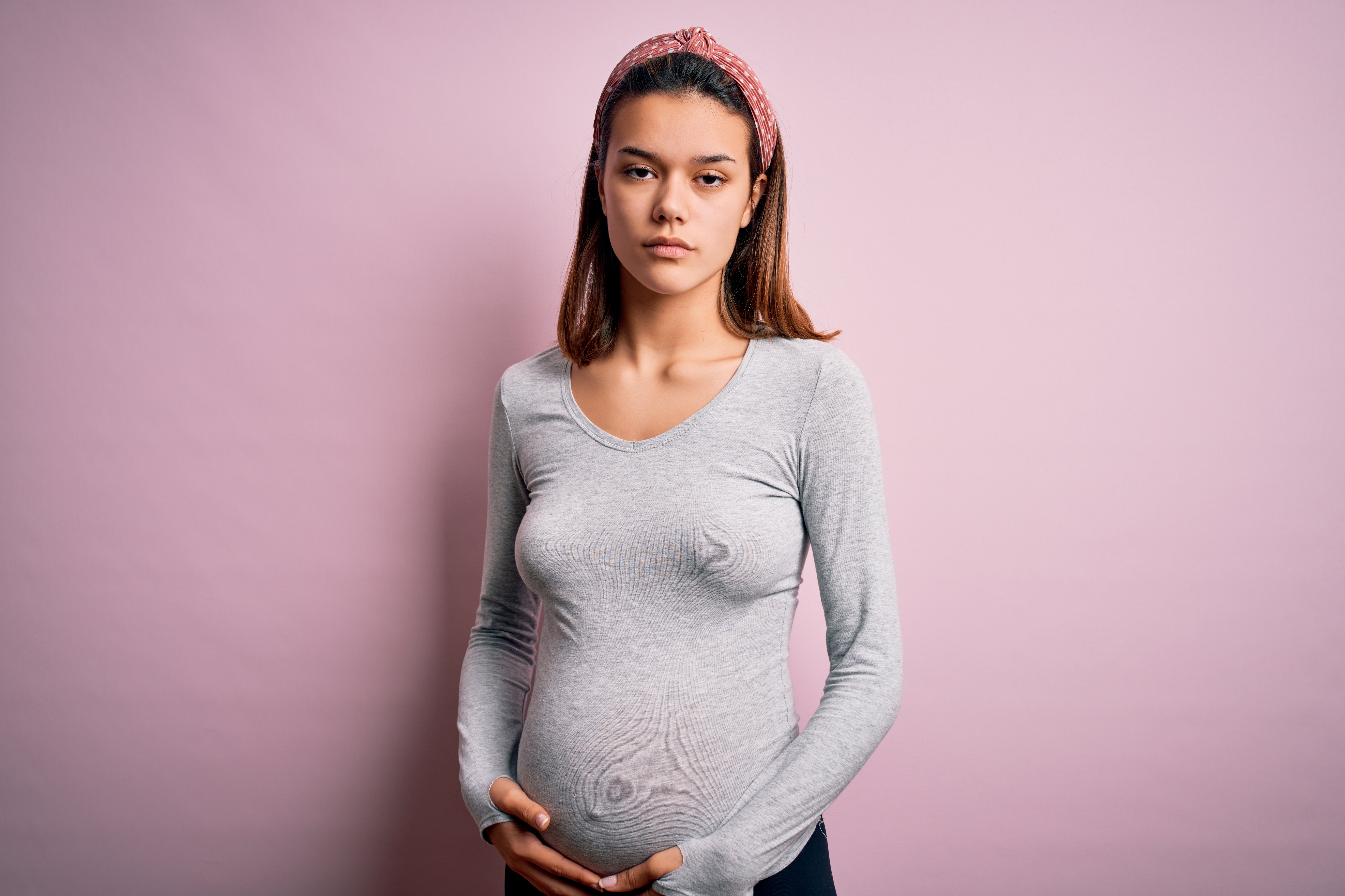 Eine hochschwangere junge Frau mit ernstem Gesichtsausdruck steht vor einer rosafarbenen Wand und hält sich den Bauch.