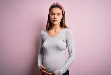 Eine hochschwangere junge Frau mit ernstem Gesichtsausdruck steht vor einer rosafarbenen Wand und hält sich den Bauch.
