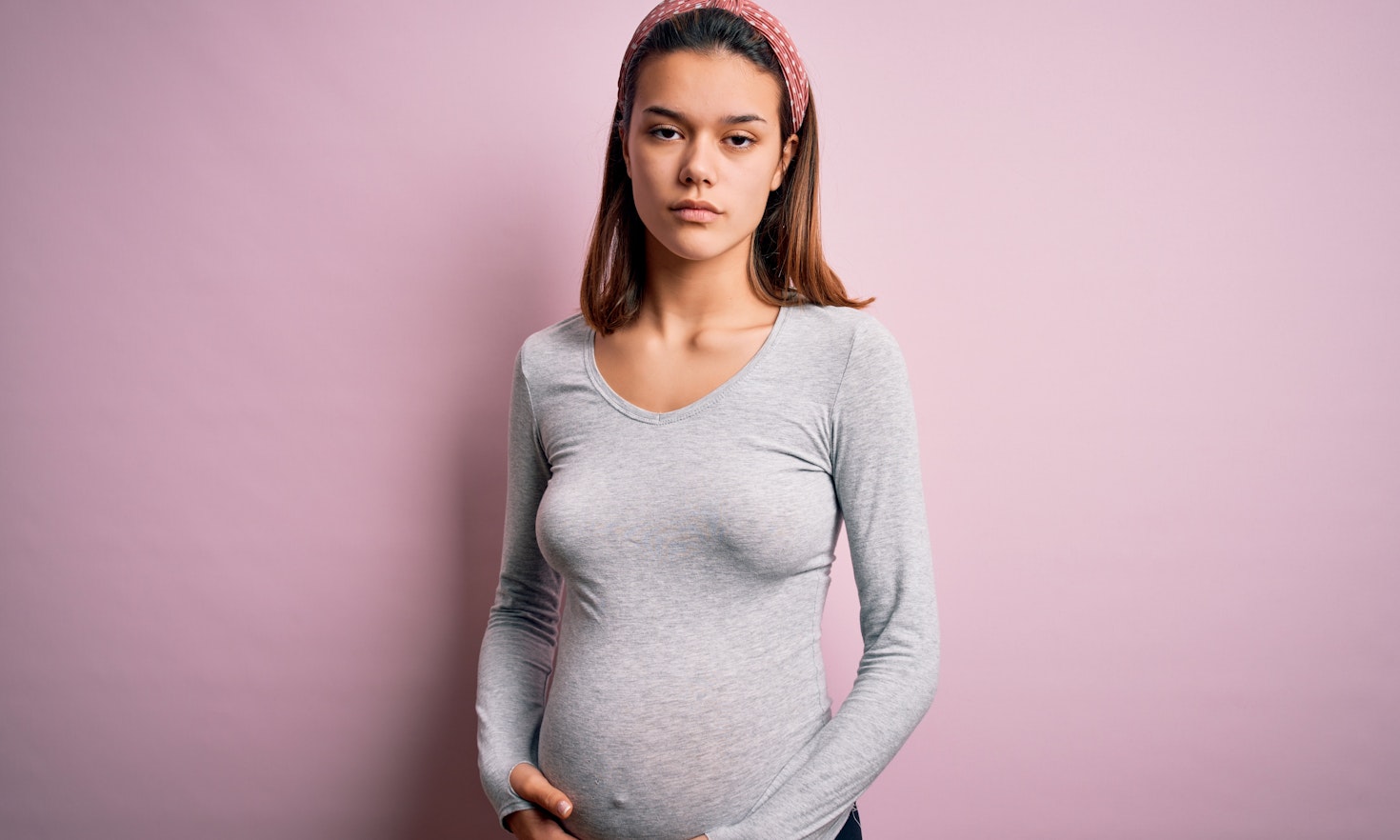 Eine hochschwangere junge Frau mit ernstem Gesichtsausdruck steht vor einer rosafarbenen Wand und hält sich den Bauch.