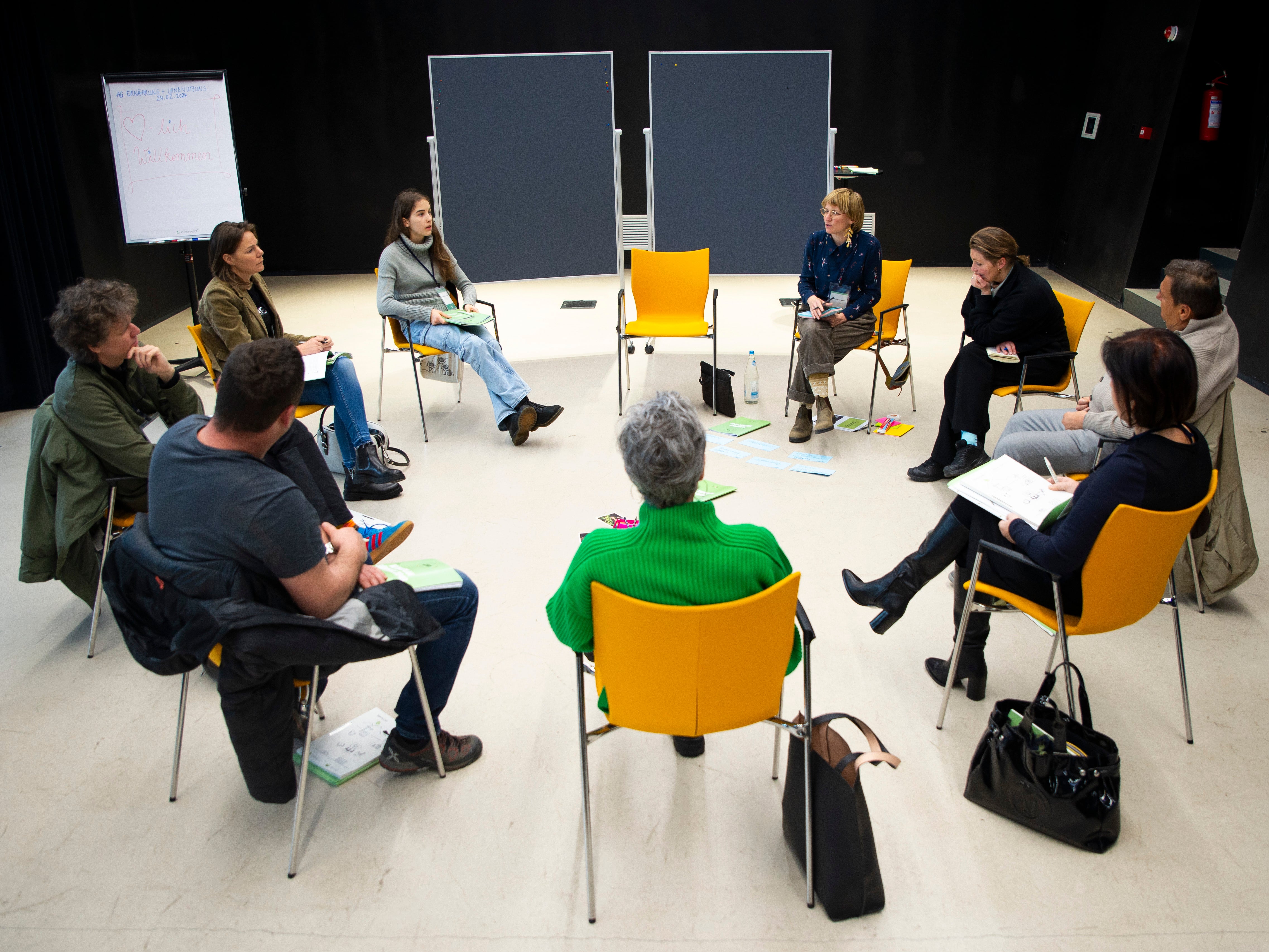 Nine climate citizens' assembly working group members are sitting on yellow chairs in a circle. Some put their bags on the floor, and there are coloured index cards on the floor. The well-lit room has black walls, and two blackboards can be seen in the background.
