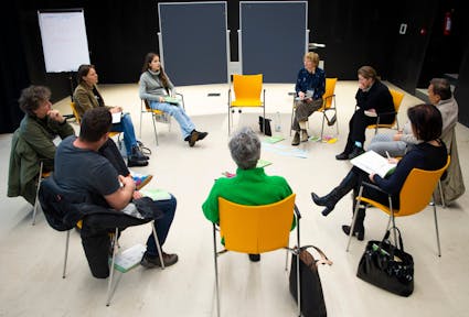 Nine climate citizens' assembly working group members are sitting on yellow chairs in a circle. Some put their bags on the floor, and there are coloured index cards on the floor. The well-lit room has black walls, and two blackboards can be seen in the background.
