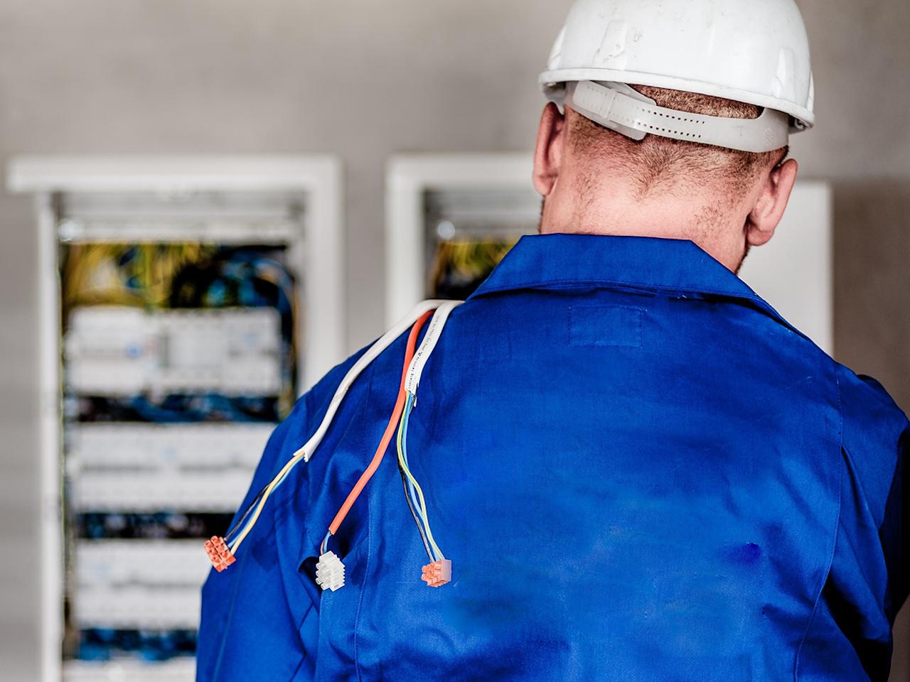 Un elettricista di spalle, con elmetto protettivo, di fronte a un pannello elettrico